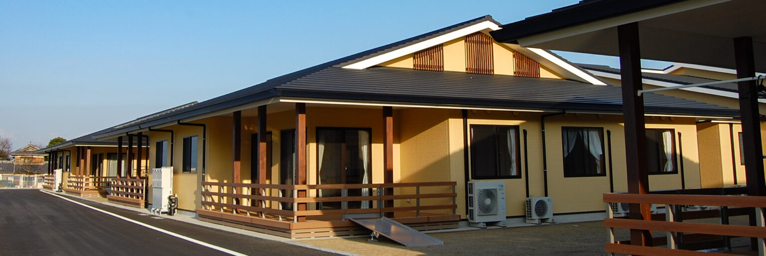 木造平屋建ての介護施設外観。温かみのある黄色い壁と木製の庇が特徴的。